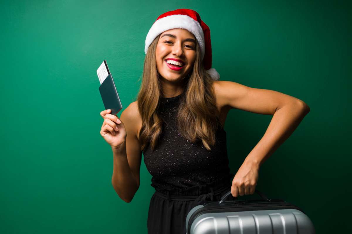 ragazza sorridente con cappello di Babbo Natale con biglietto e valigia in mano parte per le vacanze natalizie